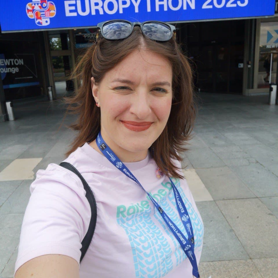 Selfie of Ester Beltrami in front of the Congress Center in Prague during Europython 2023. Ester is smiling at the camera, wearing a pink t-shirt and a lanyard with her attendee badge, with a large blue sign reading 'EUROPYTHON 2023' visible above her.